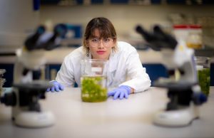 Radha Gomez wears a lab coat and purple nitrile gloves. She is seated at a lab table peering over a jar containing plant matter looking into the camera through her round rimmed glasses. anta Fe College series taken in the X Building labs on Aug. 10, 2022 in Gainesville.