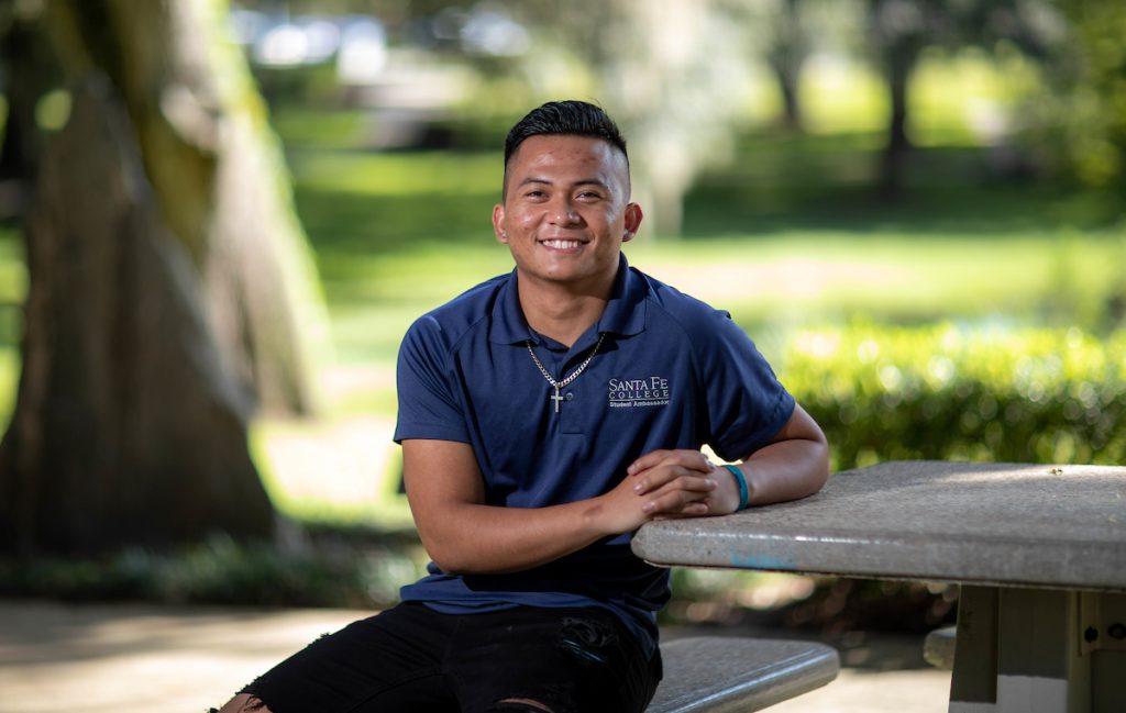 Saint of Santa Fe College John Bescoro is a Student Ambassador poses for a photo on Sept. 22, 2021, sitting at a picnic table with green treelined background Gainesville , FL.