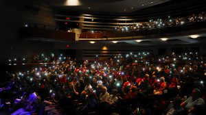 Audience in the Jackson N. Sasser Fine Arts Hall at Santa Fe College sit in the dark with lights illuminated like fireflies coming from cell phones in the audience.