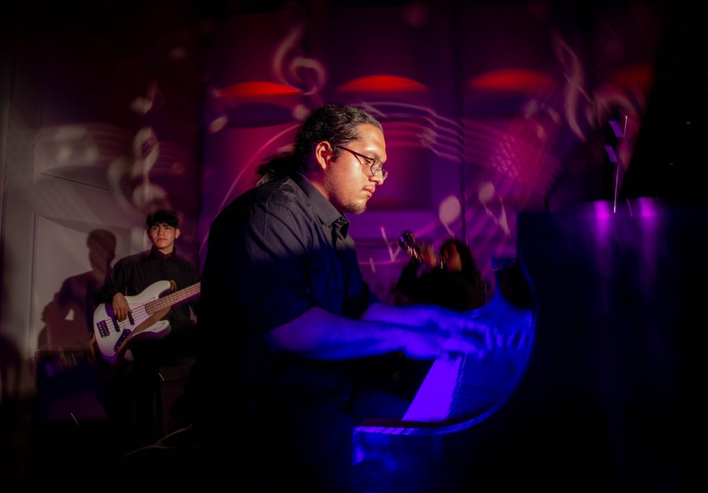 Man sits in dark room illuminated in blue and red, playing hte piano.