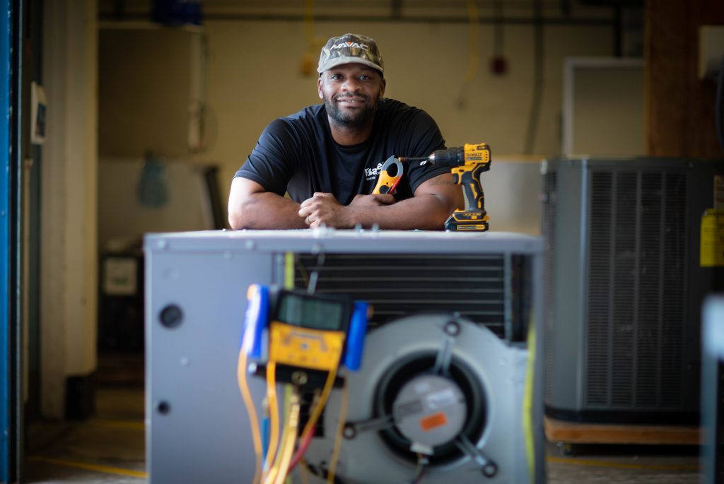 Recent graduate, Jeremiah Neal kneels behind an air conditioning unit with arms resting on top of the unit as he holds a battery operated drill. Wearing a ball cap he looks straight into the camera.