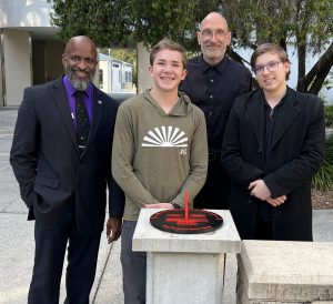 four people posing in front of a red sun dial on a stand