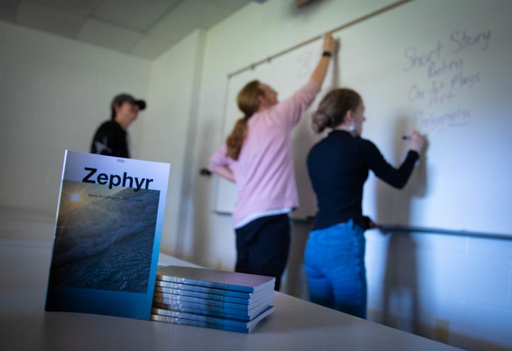 a book with the word "Zephyr" sits on a table, in the background two people in in a black shirt and one in a pink shirt write on a white board while one person in a black shirt and hat stands to the side and watches