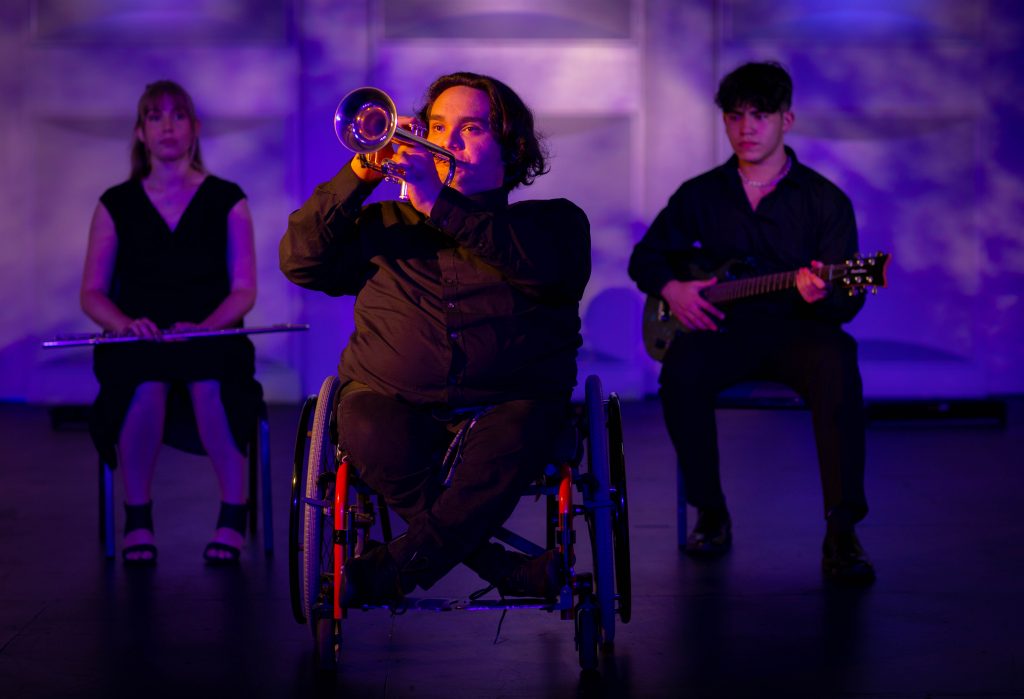 a man wearing all black in a wheelchair playing a trumpet in the background are two people a man wearing all black holding a guitar and a woman wearing a black holding a flute