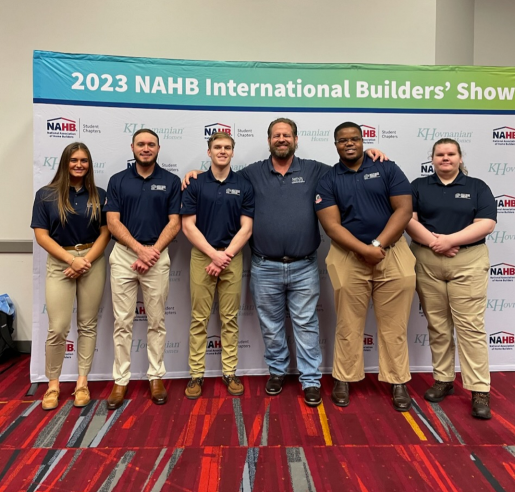 six people standing in front of a white background with blue matching polos the background reads"2023 NAHB International Builders Show"