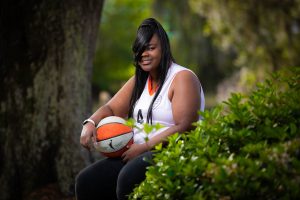 Ivory Richardson, a woman with medium brown skin and dark hair with bangs draped across her face sits with an orange and white basketball in her lap wearing a white tank top, sitting for a portrait.