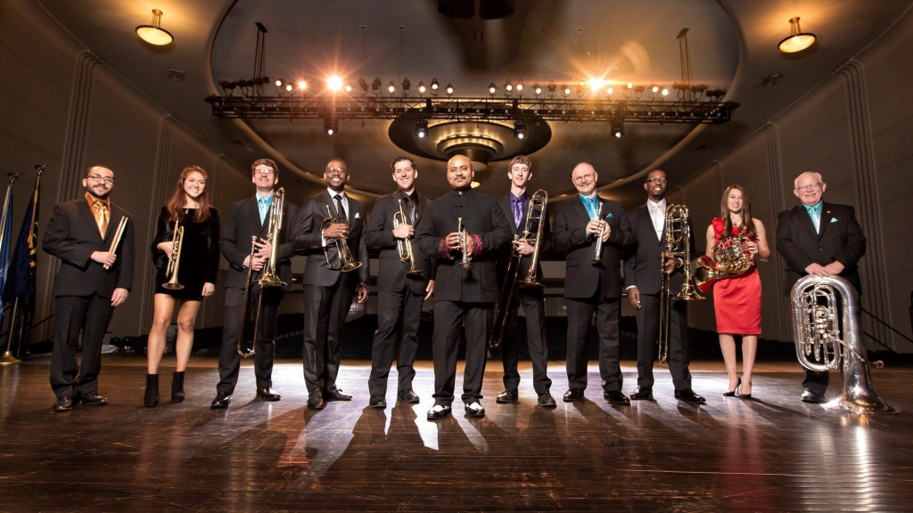 The 11 members of the Rodney Marsalis Philadelphia Big Brass Ensemble standing across a stage each holding their respective instrument. Rodney Marsalis is in the center of the group.
