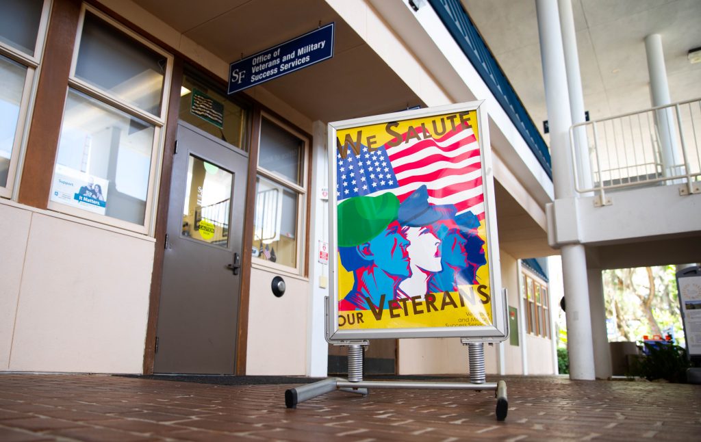 A sign with illustrations of enlisted individuals that reads "We Salute Our Veterans" on the sidewalk in front of the Santa Fe College Veterans and Military Success Services office 