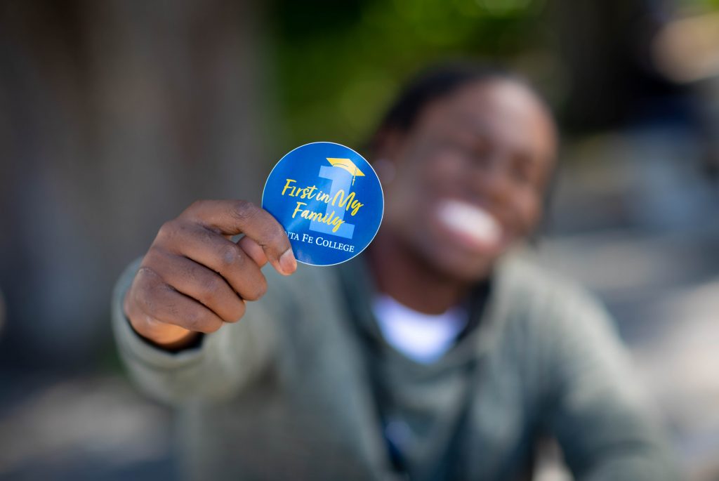 An individual holding out a button that reads "First in My Family. Santa Fe College."