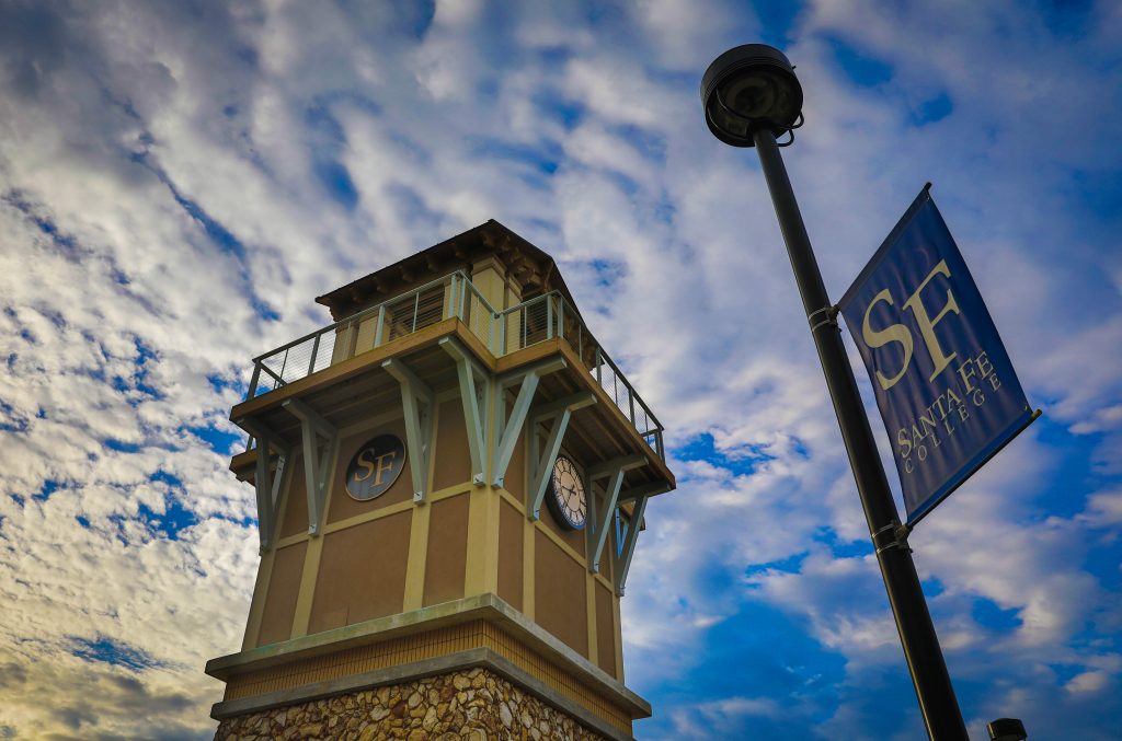 The Santa Fe College Clock Tower next to a Santa Fe College pole banner against a blue sky with clouds