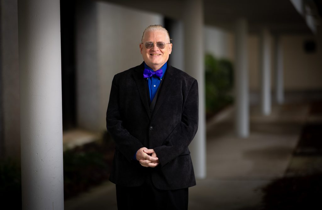 Santa Fe College Joseph Jester, Executive Assistant to the Vice President dressed in a suit and bow tie, stands with his fingers clasped in front of him in a campus walkway