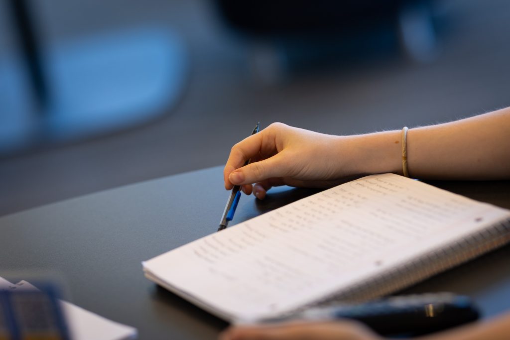 A hand holding a pen with a notebook open on a black table in front of the person.