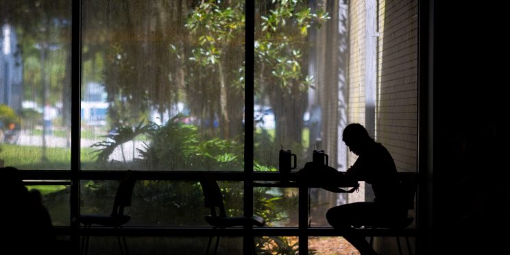 A person sitting in shadow at a table in a dark place with a bright backdrop of trees, bushes and unfocused objects outside a wide window.