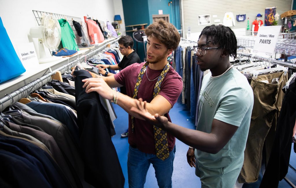 Two men browsing clothes in a clothing store, examining different garments and discussing their choices.