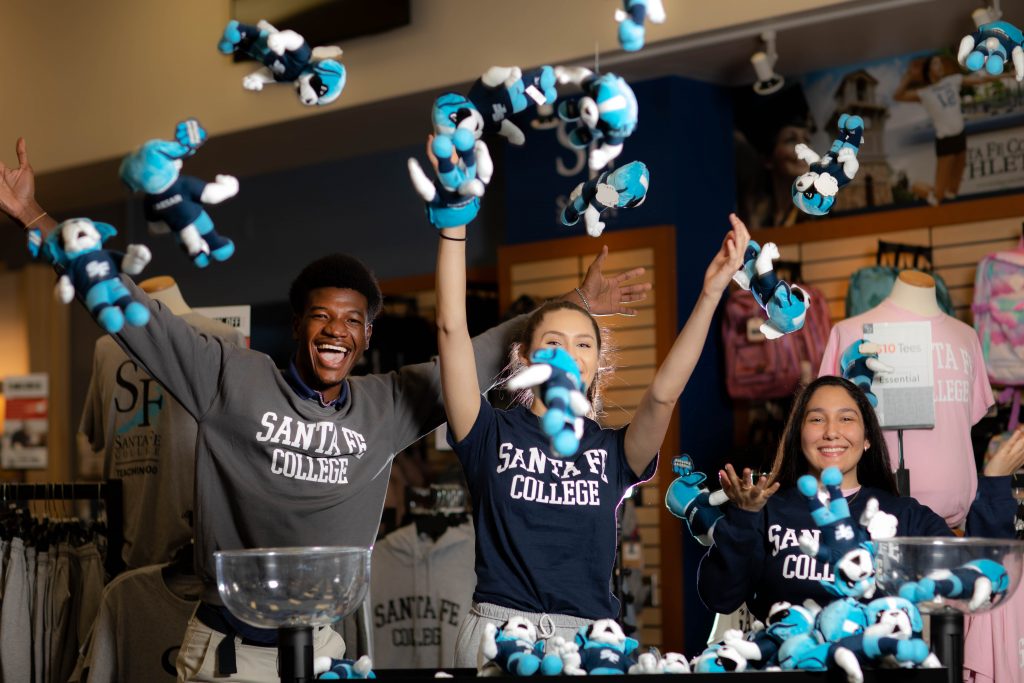 Three smiling people dressed in shirts that read "Santa Fe College" throw small Caesar Saint plush dolls into the air in the Santa Fe College Bookstore.