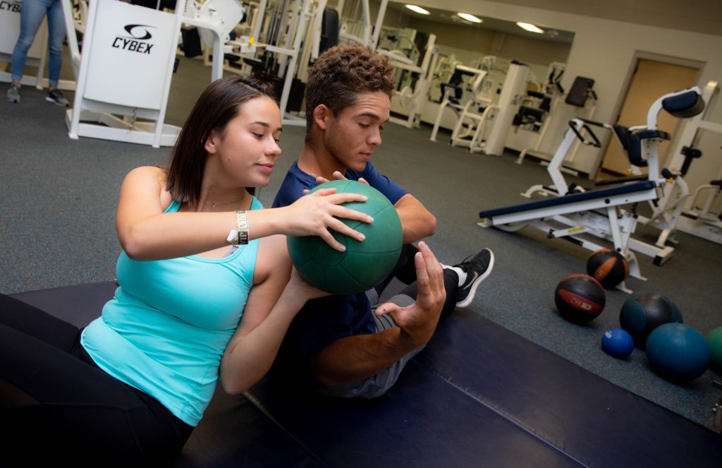 One person passes a ball over their shoulder to someone else while they sit on a floor mat with workout equipment around them.