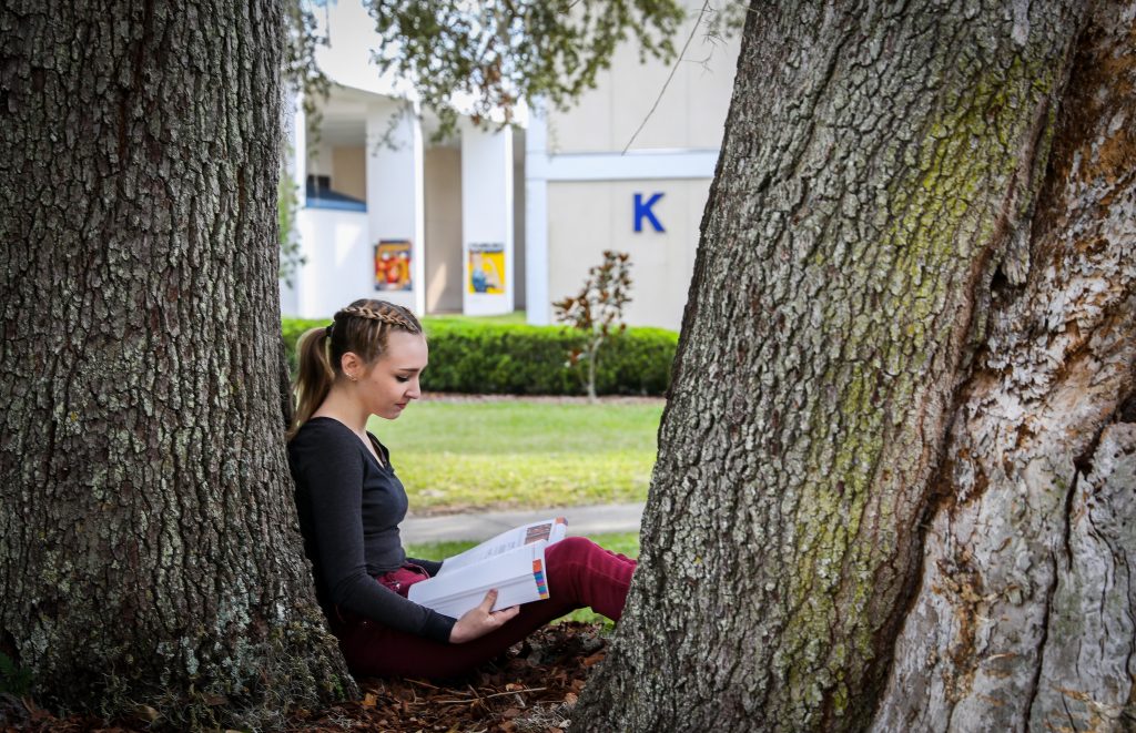 A person sits against a tree as they read a book on Santa Fe College' Northwest Campus