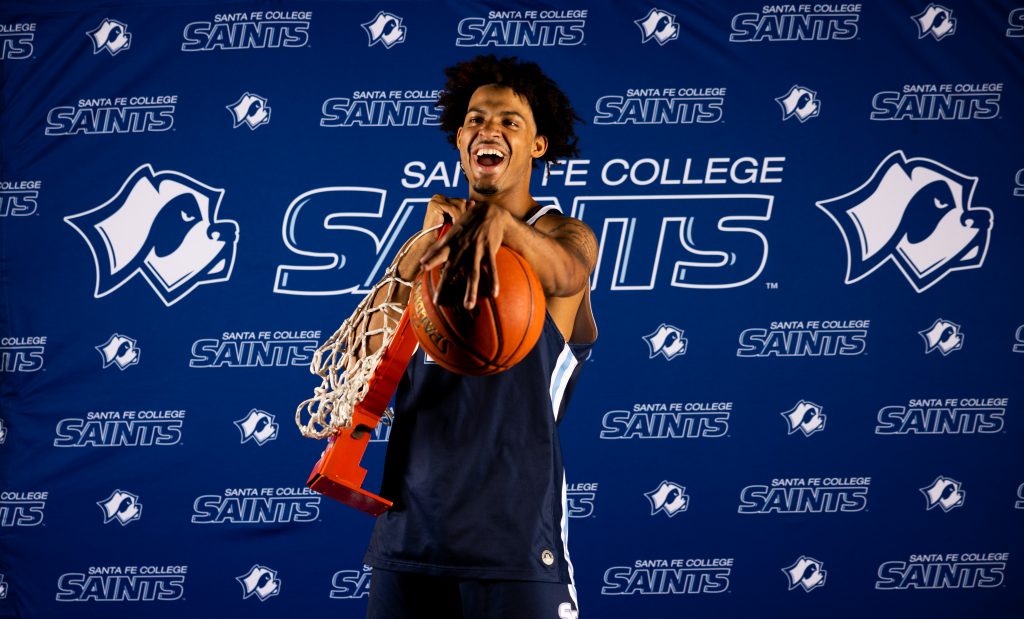 Santa Fe College Saints Men’s Basketball player Ashton Lovette laughs while holding a basketball net and ball in front of a backdrpo that reads "Santa Fe College Saints."