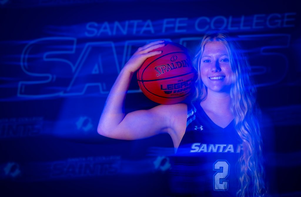 Santa Fe College Saints Women’s Basketball player Camryn Robinson smiles while holding a basketball on their shoulder.
