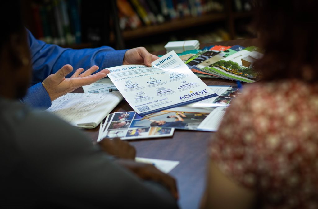 An individual is presenting an SF Achieve information flyer to two other individuals at a table in a library.