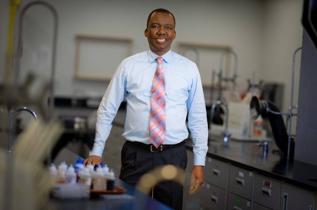 Santa Fe College chemistry professor Alpheus Mautjana, dressed in a long-sleeved shirt, tie, and slacks, stands smiling in a chemistry lab.