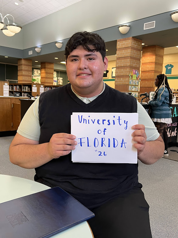 Jesus Dominguez sits smiling while holding a sign that reads" University of Florida '26."