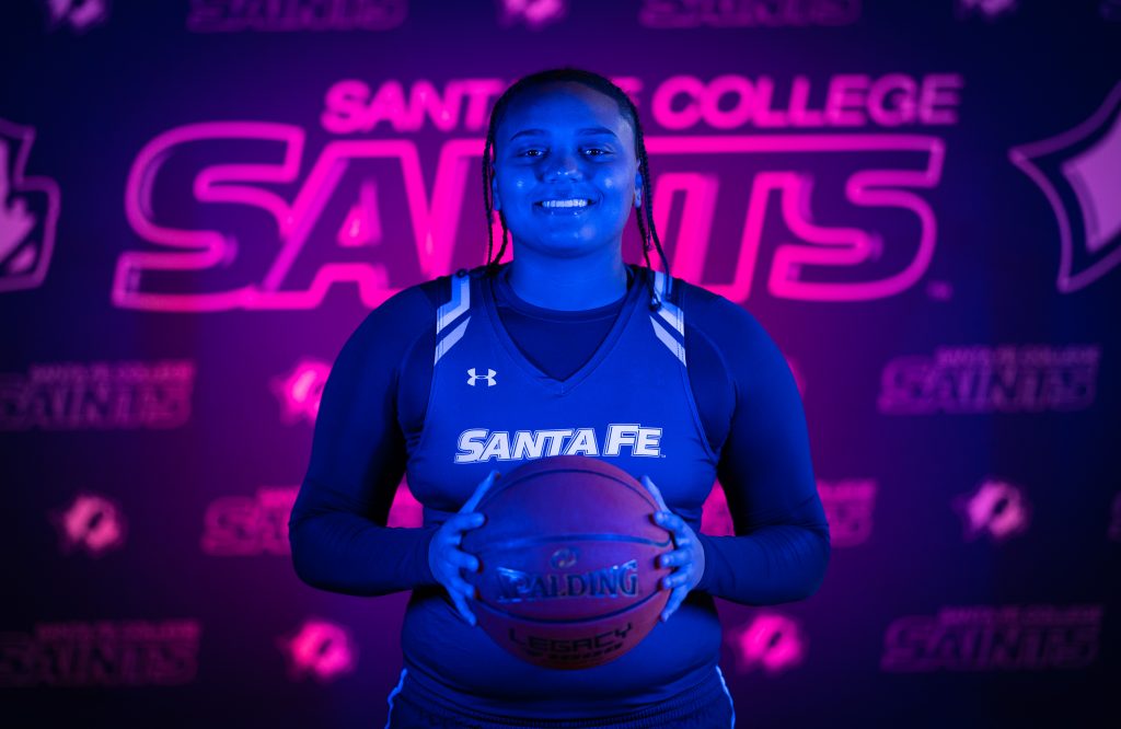 Santa Fe College Saints Women’s Basketball player Joey Delancy smiles while holding a basketball between their hands in an indoor photoshoot.