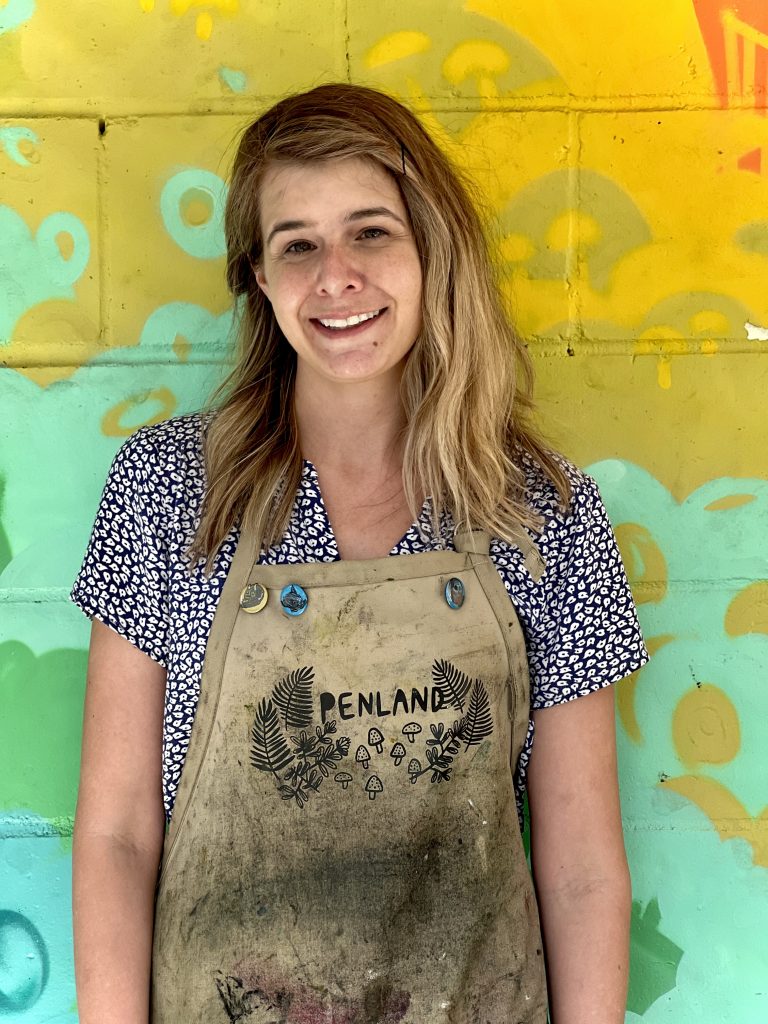 Molly Kempson poses for a photograph while standing in front of a colorful brick wall and wearing an apron.