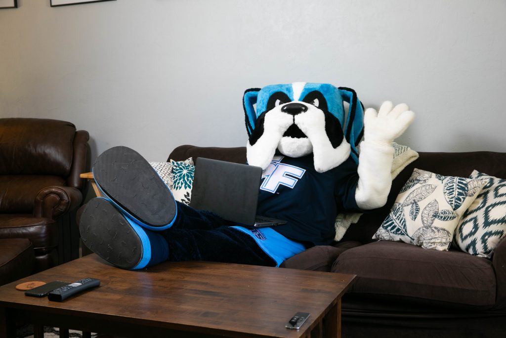 Santa Fe College mascot Caesar Saint sits on a couch waving while having a laptop on his lap.