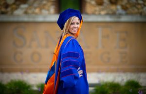 An individual poses for the camera while wearing a blue and orange graduation cap and gown.
