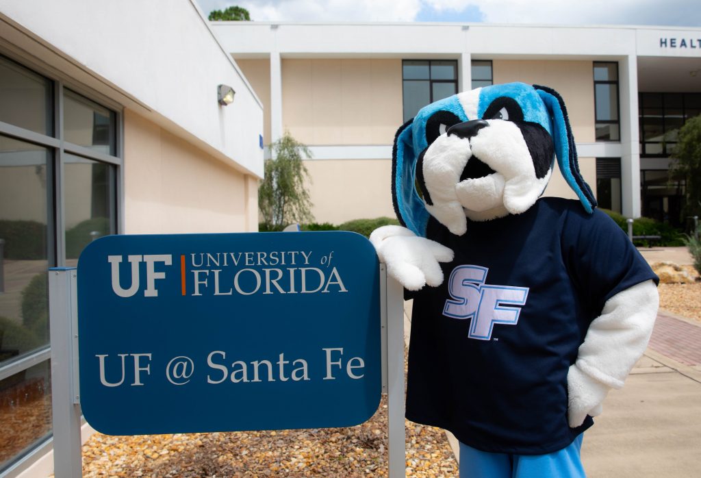 Santa Fe College mascot Caesar Saint stands outside the SF @ Santa Fe office, leaning on the office's sign.