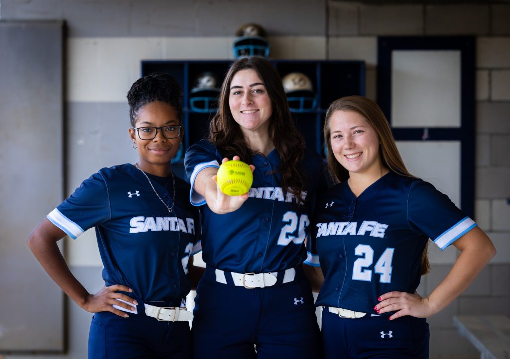 Santa Fe College Saints Softball players Jayla Clark, Mallory Forrester and Peyton Bass stand in the dugout. Mallory holds a softball up.