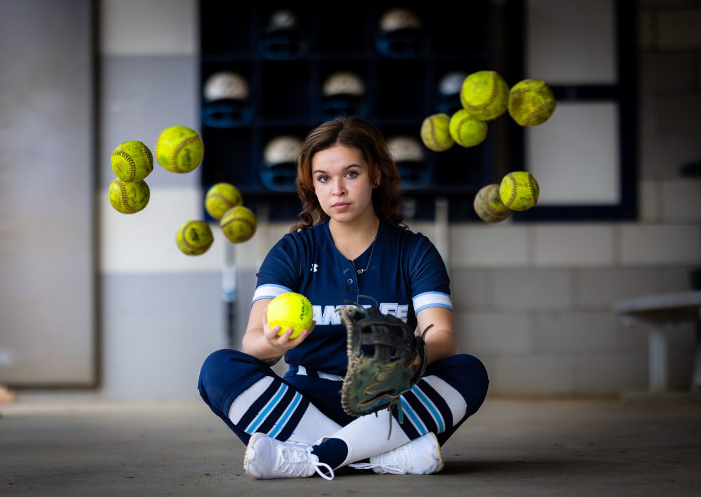 Santa Fe College Saints Softball Player Anna Burnside sits in a dugout with softballs hovering around her.