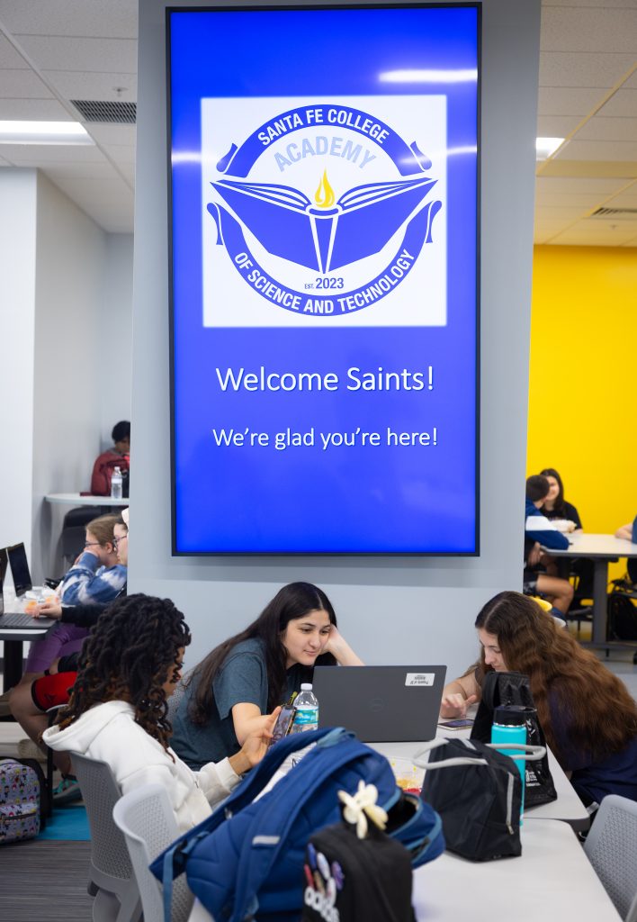 Individuals sitting under a monitor that displays the Santa Fe College Academy of Science and Technology and text that reads: "Welcome Saints! We're glad you're here!"