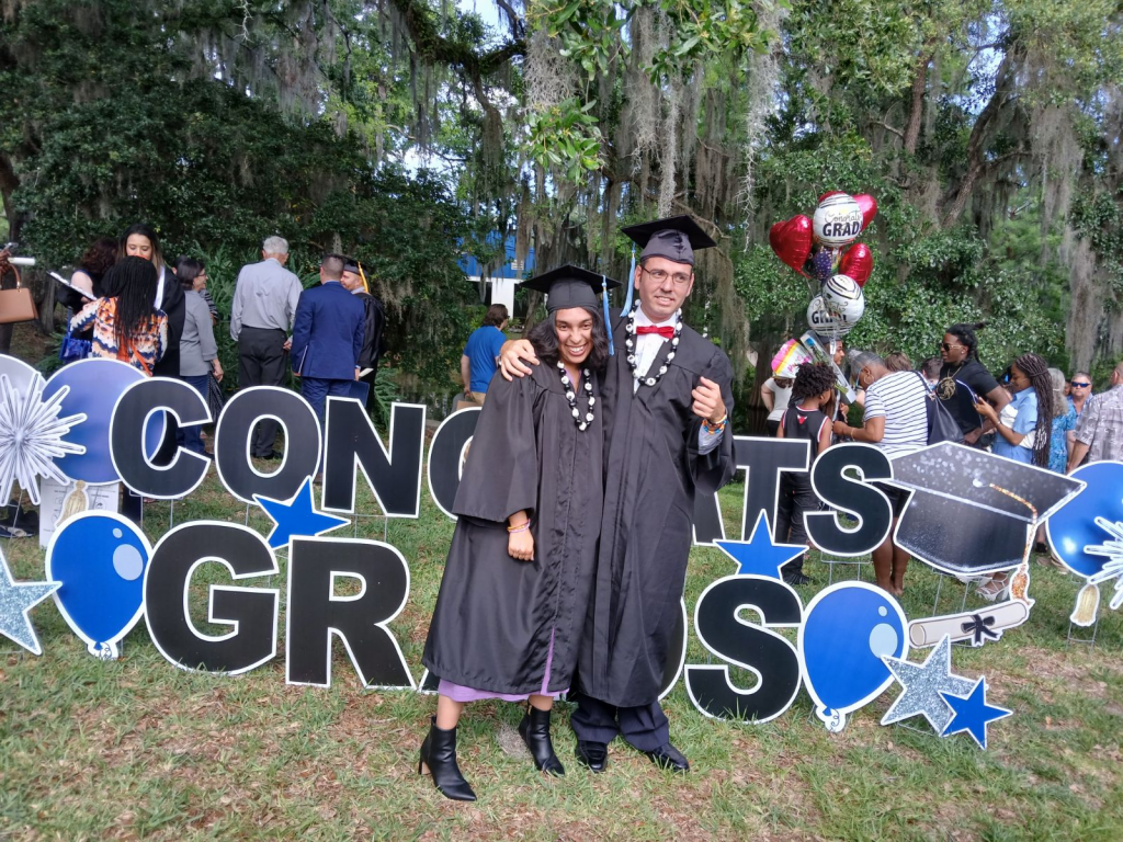 Two individuals in graduation gowns and cap standing in front of large black letters