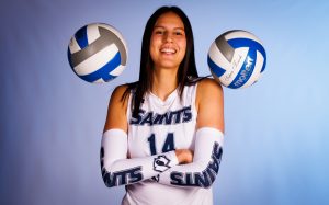 A smiling individual in a volleyball jersey with two volleyballs hovering above each shoulder.