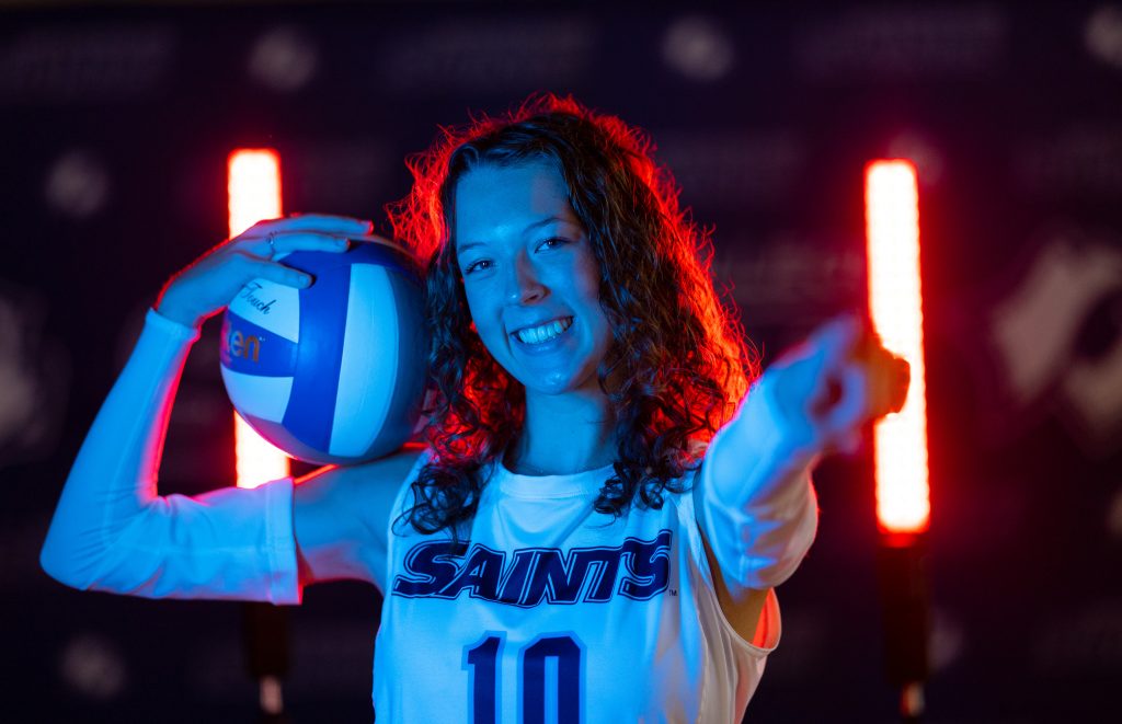 An individual holding a volleyball on their bicep points at the camera.