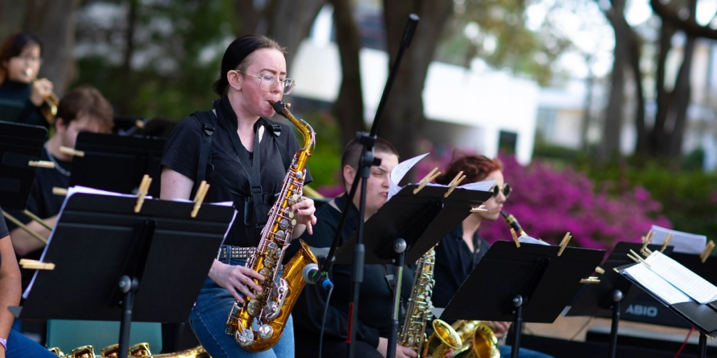 An individual playing a saxophone on an outdoor stage with other musicians in the background.